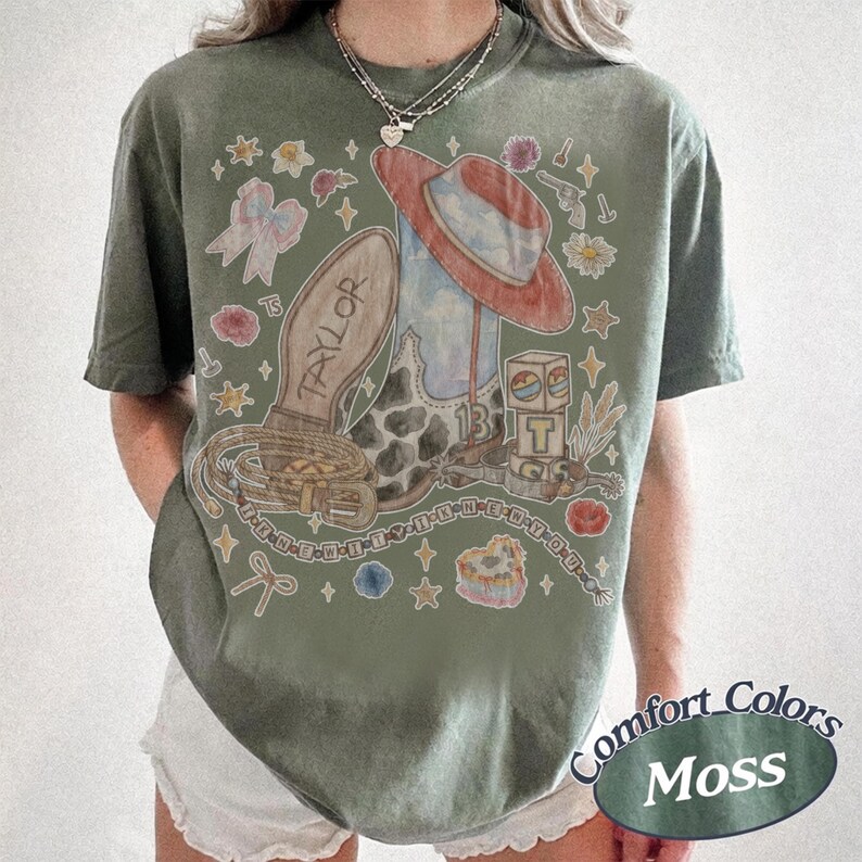 May include: A moss green short-sleeve t-shirt featuring a graphic print of a cowboy boot, a red hat, a lasso, and a block with the letter T. The text TAYLOR is printed on the boot sole. Additional illustrations include stars, flowers, and bows.