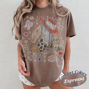 May include: An espresso-colored t-shirt featuring a graphic print of a cowboy boot, a red hat, a belt, and a block with the letter T. The design includes small illustrations of bows, stars, flowers, and the text Taylor and 13.