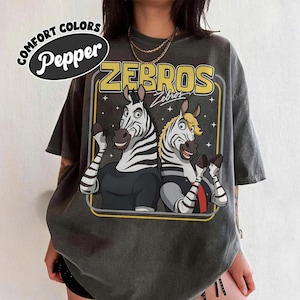 May include: Dark grey t-shirt with a cartoon graphic of two zebras and the word "ZEBROS". The shirt has a vintage, washed look. The words "COMFORT COLORS" and "Pepper" are printed on the top left.