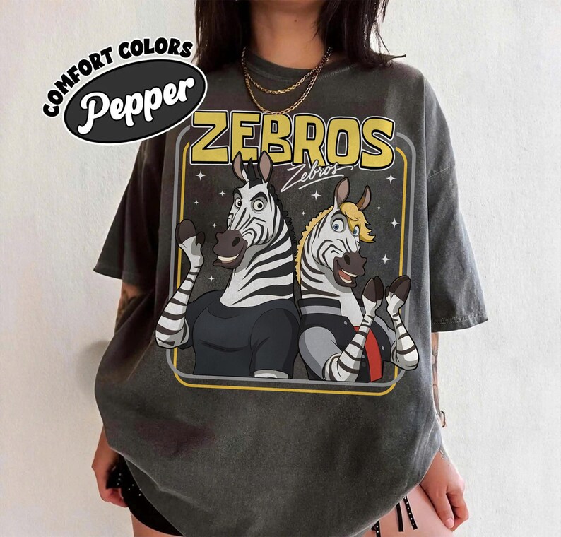 May include: Dark grey t-shirt with a cartoon graphic of two zebras and the word "ZEBROS". The shirt has a vintage, washed look. The words "COMFORT COLORS" and "Pepper" are printed on the top left.