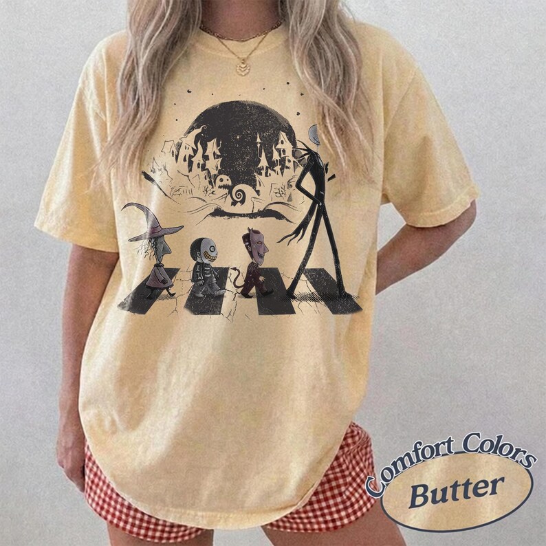 May include: A pale yellow short-sleeve t-shirt featuring a black graphic print of characters walking across a crosswalk in front of a dark moon and a spooky house silhouette. The design includes a witch, a skeleton, and a devilish figure.