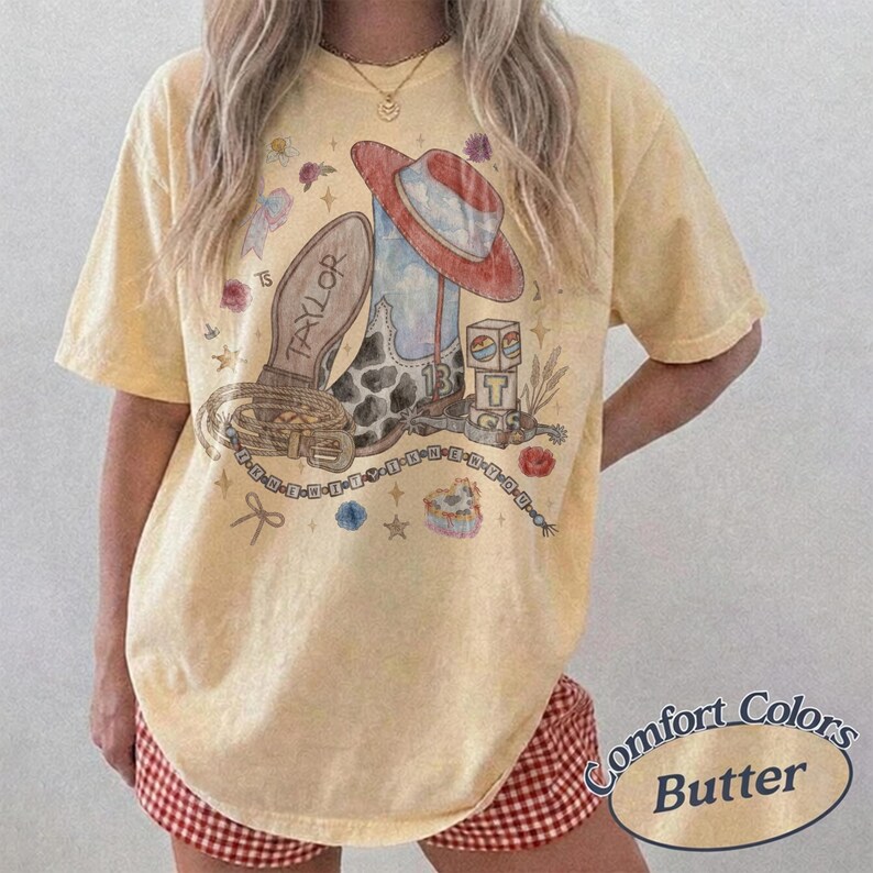 May include: A pale yellow short-sleeve t-shirt featuring a graphic print of a cowboy boot, a hat, a belt, and a block with the number 13. The word TAYLOR is printed on the boot. The design includes small stars, flowers, and a beaded chain.