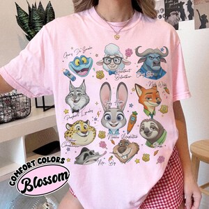 May include: A light pink t-shirt featuring cartoon characters from a movie. The shirt includes the text "COMFORT COLORS Blossom" in a circular design. The characters include a frog, sheep, buffalo, fox, rabbit, and sloth.