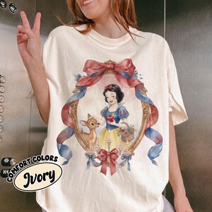 May include: Ivory-colored t-shirt featuring a framed illustration of Snow White with a deer, holding an apple and a basket of flowers. The frame is adorned with red and blue ribbons and bows. The words "COMFORT COLORS" and "Ivory" are printed on the shirt.