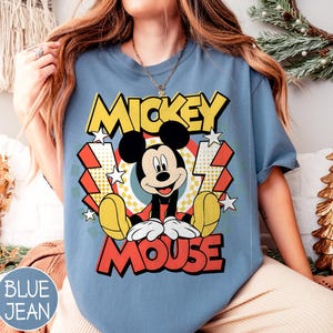 May include: Blue jean colored t-shirt featuring a graphic of Mickey Mouse. The design includes the words "Mickey Mouse" in yellow and red, with a cartoon image of Mickey. The shirt has short sleeves.