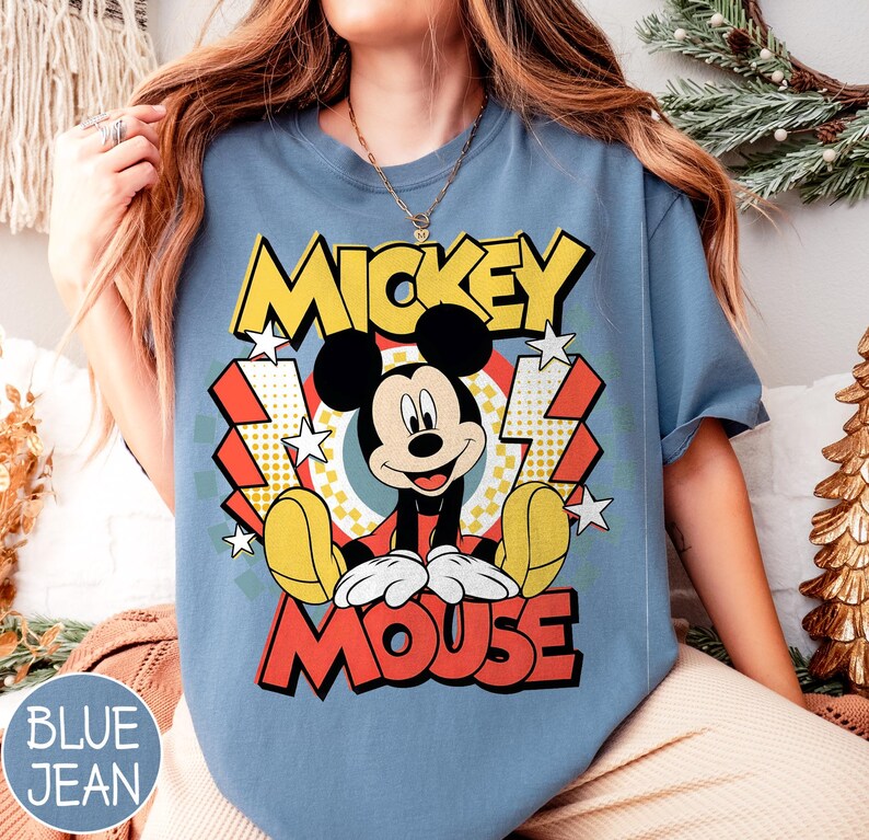 May include: Blue jean colored t-shirt featuring a graphic of Mickey Mouse. The design includes the words "Mickey Mouse" in yellow and red, with a cartoon image of Mickey. The shirt has short sleeves.