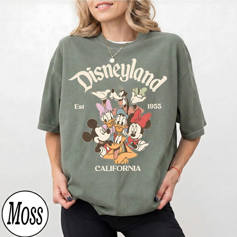Puede incluir: Camiseta verde salvia con el logotipo de Disneyland y personajes de dibujos animados. El texto "Disneyland" est&aacute; arqueado en la parte superior, con "Est 1955" debajo. La palabra "CALIFORNIA" est&aacute; en la parte inferior.