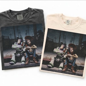 May include: Two t-shirts, one dark gray and one cream, each featuring a graphic of Buzz Lightyear and Woody from Toy Story. The characters are depicted sitting and holding bottles, set against a nighttime street scene.