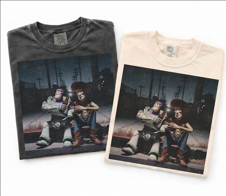 May include: Two t-shirts, one dark gray and one cream, each featuring a graphic of Buzz Lightyear and Woody from Toy Story. The characters are depicted sitting and holding bottles, set against a nighttime street scene.