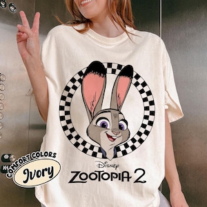 May include: Ivory-colored t-shirt featuring a cartoon image of Judy Hopps from Disney's Zootopia 2, set within a black and white checkered circle. The shirt also has the words "COMFORT COLORS" and "Disney Zootopia 2" printed on it.