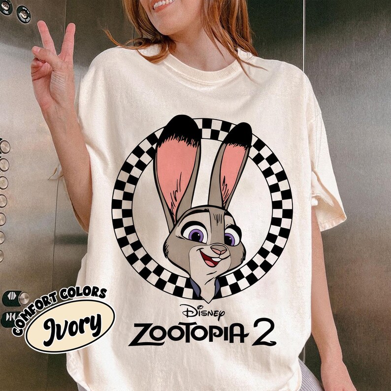 May include: Ivory-colored t-shirt featuring a cartoon image of Judy Hopps from Disney's Zootopia 2, set within a black and white checkered circle. The shirt also has the words "COMFORT COLORS" and "Disney Zootopia 2" printed on it.
