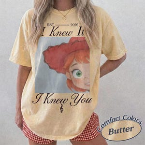 May include: A pale yellow short-sleeve t-shirt featuring a graphic of a red-haired character wearing a red hat. The text reads EST 2026 I Knew It and I Knew You with a musical note symbol. The shirt has a vintage-style washed appearance.