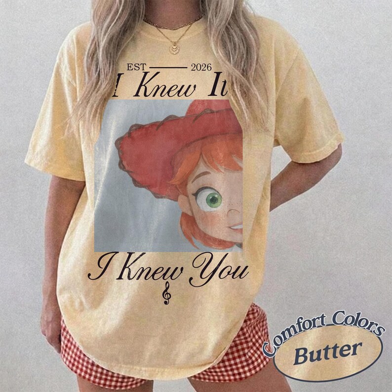 May include: A pale yellow short-sleeve t-shirt featuring a graphic of a red-haired character wearing a red hat. The text reads EST 2026 I Knew It and I Knew You with a musical note symbol. The shirt has a vintage-style washed appearance.