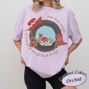 K&ouml;nnte beinhalten: Ein hellviolettes Kurzarm-T-Shirt mit einer Grafik, die einen Reifen zeigt, der Zeichentrickfiguren, eine Gitarre und einen roten Cowboystiefel umrahmt. Der Text lautet Love has ways of bringing things back to life. Das Shirt hat einen Rundhalsausschnitt.