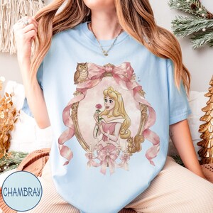 May include: Light blue t-shirt featuring a watercolor illustration of a princess holding a rose, framed by a pink bow and ornate gold detailing. An owl and squirrel are also depicted. The text "CHAMBRAY" is printed on the bottom left.
