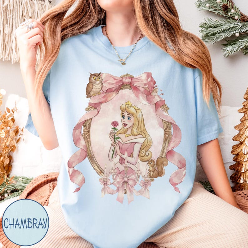 May include: Light blue t-shirt featuring a watercolor illustration of a princess holding a rose, framed by a pink bow and ornate gold detailing. An owl and squirrel are also depicted. The text "CHAMBRAY" is printed on the bottom left.