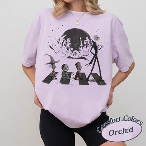 May include: A light purple t-shirt featuring a black graphic print of characters walking across a crosswalk. The design includes a tall skeletal figure, three smaller figures, and a dark moonlit background with a silhouette of a town.