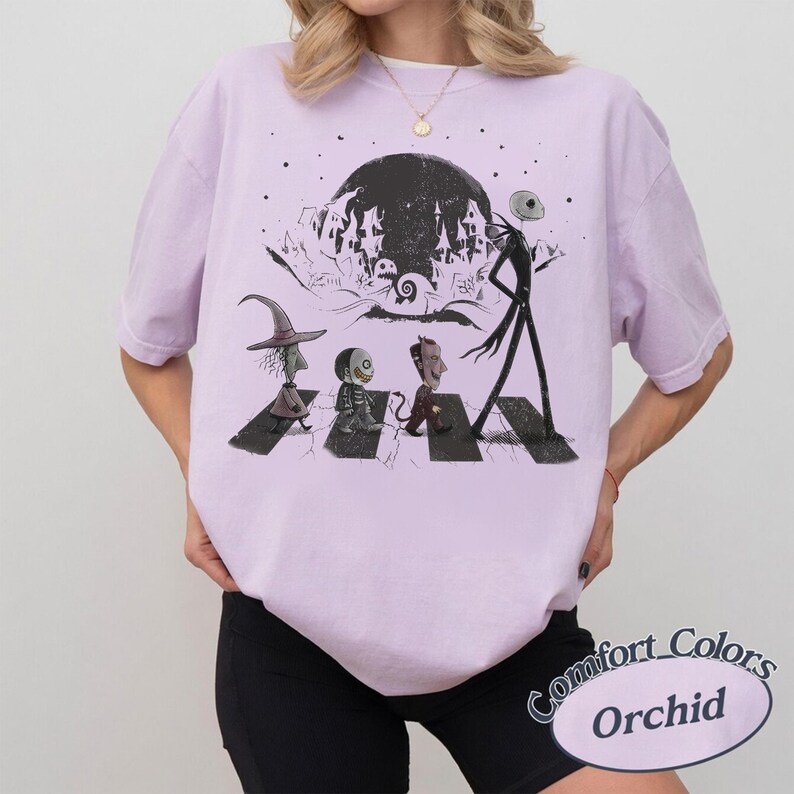 May include: A light purple t-shirt featuring a black graphic print of characters walking across a crosswalk. The design includes a tall skeletal figure, three smaller figures, and a dark moonlit background with a silhouette of a town.