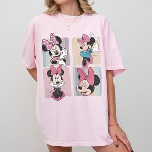 Op de afbeelding: Een lichtroze t-shirt met een afbeelding van Minnie Mouse in vier verschillende poses. Minnie Mouse wordt afgebeeld met haar zwarte oren, roze strik en verschillende outfits. Het ontwerp is geplaatst tegen een pastelkleurige achtergrond.