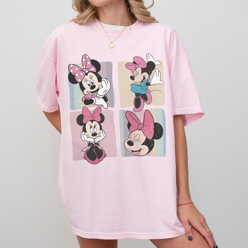 Op de afbeelding: Een lichtroze t-shirt met een afbeelding van Minnie Mouse in vier verschillende poses. Minnie Mouse wordt afgebeeld met haar zwarte oren, roze strik en verschillende outfits. Het ontwerp is geplaatst tegen een pastelkleurige achtergrond.