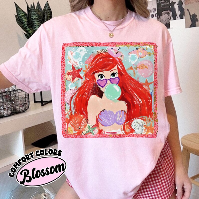 May include: Pink Comfort Colors t-shirt with a graphic of a mermaid wearing heart-shaped sunglasses and blowing a bubble. The graphic is surrounded by seashells and starfish. The word "Blossom" is printed on the bottom of the shirt.