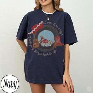 K&ouml;nnte beinhalten: Ein marineblaues Kurzarm-T-Shirt mit einer Grafik eines Reifens, der eine Gitarre, einen roten Cowboyhut und einen roten Cowboystiefel umrahmt. Der Text lautet, Love has ways of bringing things back to life, mit Zeichentrickfiguren im Reifen.