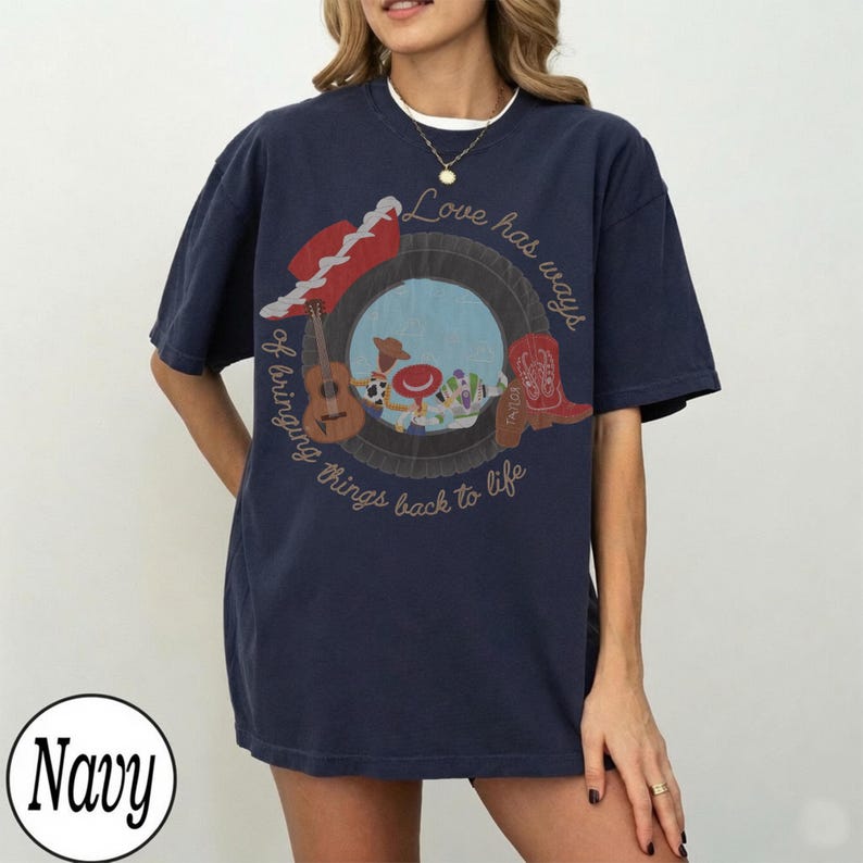 K&ouml;nnte beinhalten: Ein marineblaues Kurzarm-T-Shirt mit einer Grafik eines Reifens, der eine Gitarre, einen roten Cowboyhut und einen roten Cowboystiefel umrahmt. Der Text lautet, Love has ways of bringing things back to life, mit Zeichentrickfiguren im Reifen.