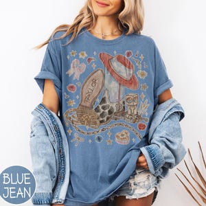 May include: A blue short-sleeve t-shirt featuring a graphic print of a cowboy boot with the name Taylor, a red hat, a belt, a block with the letter T, and various small icons including stars, flowers, and bows scattered across the front.