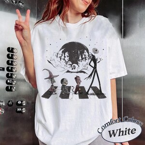 May include: A white short-sleeve t-shirt featuring a black and white graphic print of characters from The Nightmare Before Christmas walking across a crosswalk, with a large moon and a spooky town silhouette in the background.