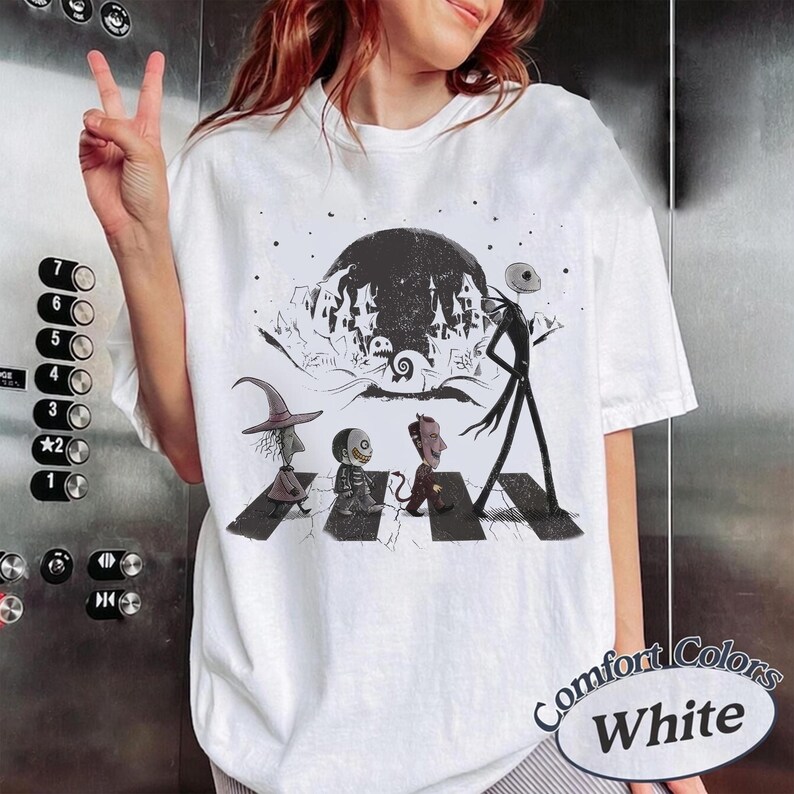 May include: A white short-sleeve t-shirt featuring a black and white graphic print of characters from The Nightmare Before Christmas walking across a crosswalk, with a large moon and a spooky town silhouette in the background.
