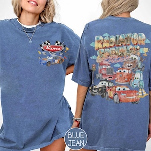 May include: A blue denim-colored t-shirt featuring a small graphic on the front chest of a blue race car with the text Name and checkered flags. The back displays a large graphic of Radiator Springs with Lightning McQueen and other cars.
