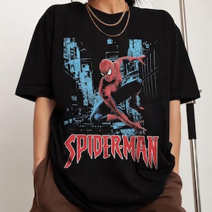May include: Black t-shirt featuring a graphic of Spider-Man in a dynamic pose against a blue cityscape backdrop. The word "SPIDERMAN" is printed in red, stylized lettering across the bottom of the shirt. The shirt has short sleeves.