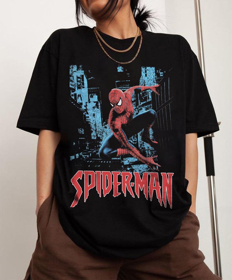 May include: Black t-shirt featuring a graphic of Spider-Man in a dynamic pose against a blue cityscape backdrop. The word "SPIDERMAN" is printed in red, stylized lettering across the bottom of the shirt. The shirt has short sleeves.