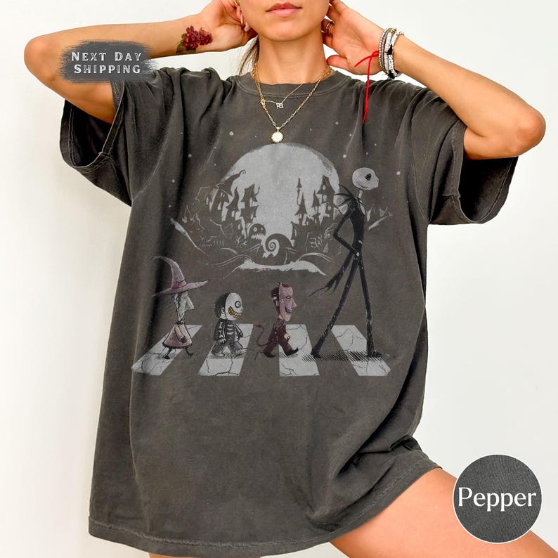 May include: A charcoal gray short-sleeve t-shirt featuring a graphic print of four characters walking across a crosswalk. The design includes a large moon, a spooky house silhouette, and the characters Lock, Shock, Barrel, and Jack Skellington.