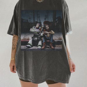 May include: Dark gray t-shirt featuring a graphic of Buzz Lightyear and Woody from Toy Story, sitting and holding bottles. The image has a vintage, distressed look. The shirt is oversized and has short sleeves.