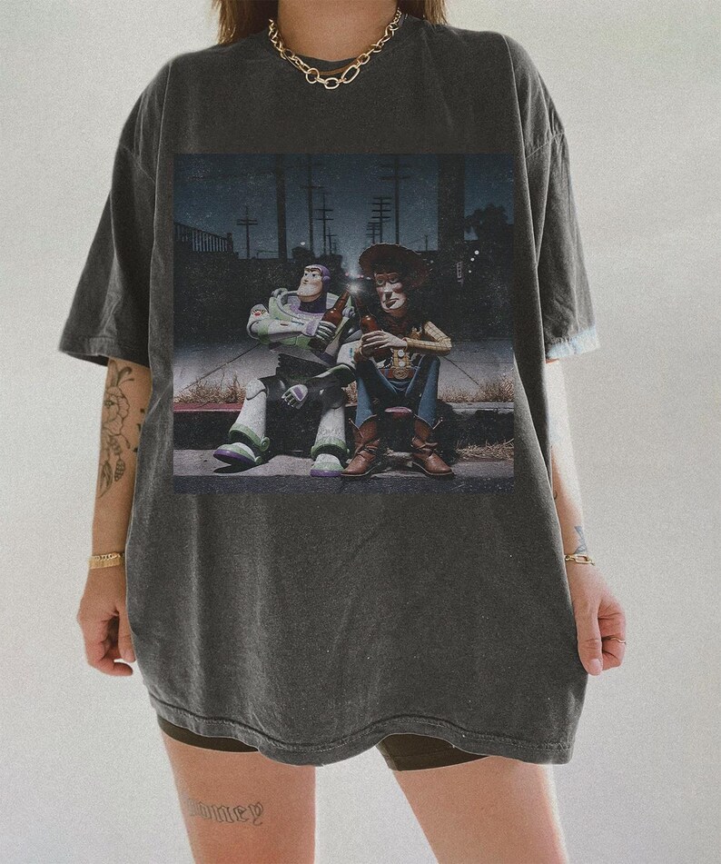 May include: Dark gray t-shirt featuring a graphic of Buzz Lightyear and Woody from Toy Story, sitting and holding bottles. The image has a vintage, distressed look. The shirt is oversized and has short sleeves.