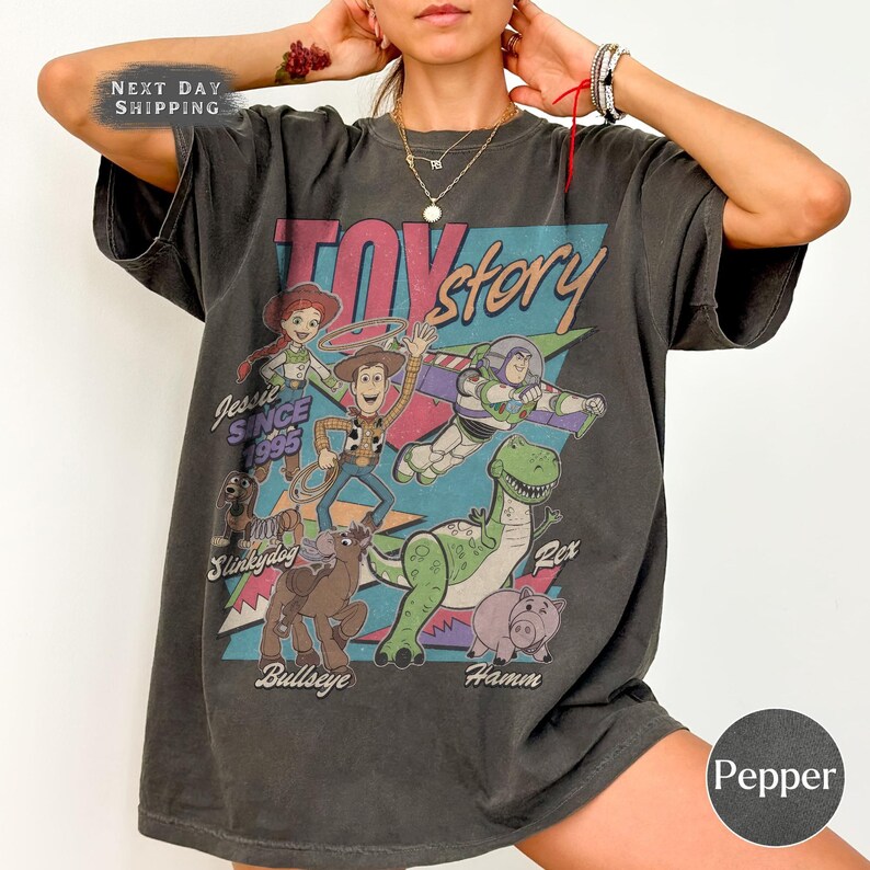May include: A charcoal grey t-shirt with a vibrant graphic of characters from the film "Toy Story." The design includes Woody, Buzz Lightyear, Jessie, Rex, Hamm, Slinky Dog, and Bullseye, with the text "Toy Story" and "Since 1995."