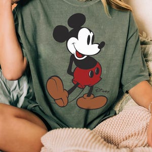 May include: An olive green t-shirt featuring a full-body graphic of Mickey Mouse. Mickey is depicted in his classic attire: black ears and body, white face, red shorts, and brown shoes. The Disney logo is printed in black near the bottom right.