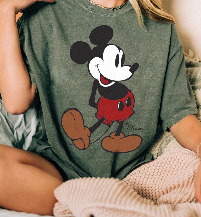 May include: An olive green t-shirt featuring a full-body graphic of Mickey Mouse. Mickey is depicted in his classic attire: black ears and body, white face, red shorts, and brown shoes. The Disney logo is printed in black near the bottom right.