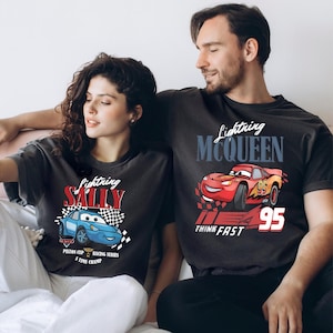 May include: Two charcoal grey t-shirts featuring Lightning McQueen and Sally Carrera from the Cars film. The Lightning McQueen shirt has a red race car graphic with the number 95. The Sally shirt has a blue race car graphic. Both shirts have text.