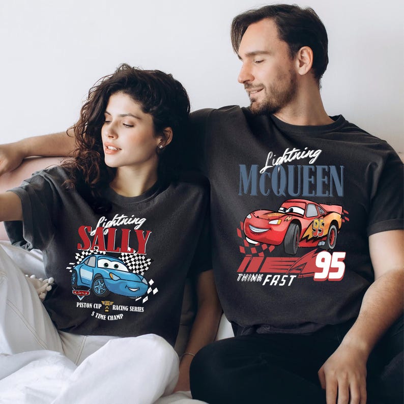 May include: Two charcoal grey t-shirts featuring Lightning McQueen and Sally Carrera from the Cars film. The Lightning McQueen shirt has a red race car graphic with the number 95. The Sally shirt has a blue race car graphic. Both shirts have text.