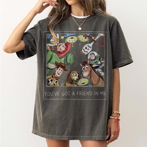 May include: Dark gray t-shirt featuring a rectangular graphic of Toy Story characters, including Woody, Buzz Lightyear, and Rex. The text "YOU'VE GOT A FRIEND IN ME" is printed below the graphic. The shirt has short sleeves.