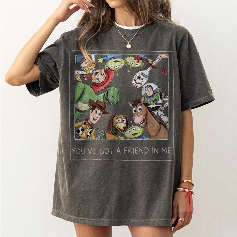 May include: Dark gray t-shirt featuring a rectangular graphic of Toy Story characters, including Woody, Buzz Lightyear, and Rex. The text "YOU'VE GOT A FRIEND IN ME" is printed below the graphic. The shirt has short sleeves.
