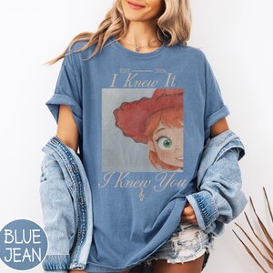 May include: A blue short-sleeve t-shirt featuring a graphic print of a cartoon character with red hair and a red hat. The text reads EST 2026 I Knew It I Knew You with a small treble clef symbol. The shirt is worn with a denim jacket and denim shorts.