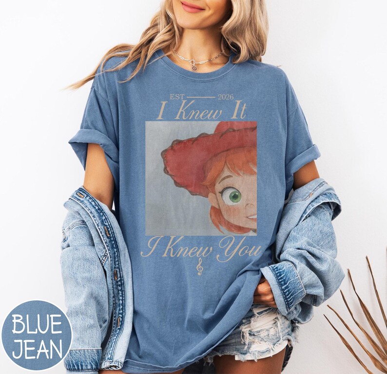 May include: A blue short-sleeve t-shirt featuring a graphic print of a cartoon character with red hair and a red hat. The text reads EST 2026 I Knew It I Knew You with a small treble clef symbol. The shirt is worn with a denim jacket and denim shorts.