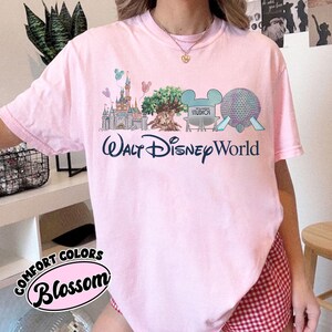 May include: A pale pink t-shirt featuring a graphic of Walt Disney World attractions, such as a castle, a tree, and Spaceship Earth. The words "Walt Disney World" are in dark blue script. The shirt also has the words "Comfort Colors" and "Blossom".