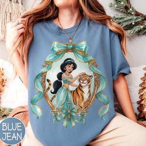 May include: Blue jean colored t-shirt featuring a framed illustration of a princess and a tiger. The illustration is framed with teal ribbons and gold accents. The text "BLUE JEAN" is in a blue circle in the lower left corner.