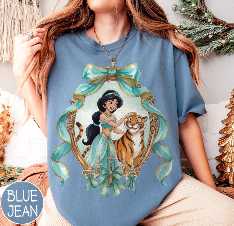 May include: Blue jean colored t-shirt featuring a framed illustration of a princess and a tiger. The illustration is framed with teal ribbons and gold accents. The text "BLUE JEAN" is in a blue circle in the lower left corner.
