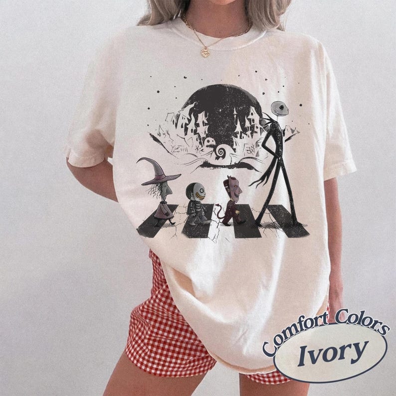 May include: An ivory short-sleeve t-shirt featuring a graphic print of four characters walking across a crosswalk. The design includes a large moon, a silhouette of a town, and the characters Lock, Shock, Barrel, and Jack Skellington.