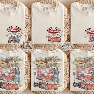 May include: Six ivory t-shirts featuring Disney Cars graphics. The top row shows individual characters with a checkered flag and a red banner labeled Name. The bottom row displays a large Radiator Springs scene with multiple characters and text.